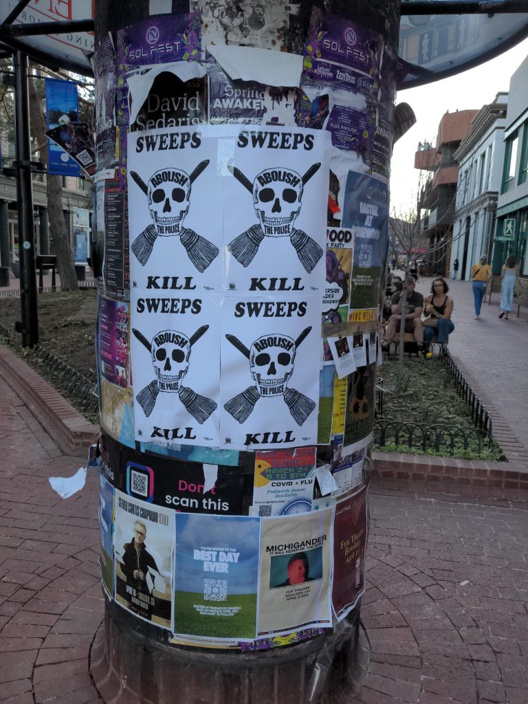 A pillar covered in posters. There are four posts in focus that say "Sweeps Kill: Abolish Police" in a drawing of a skull with brooms behind it.